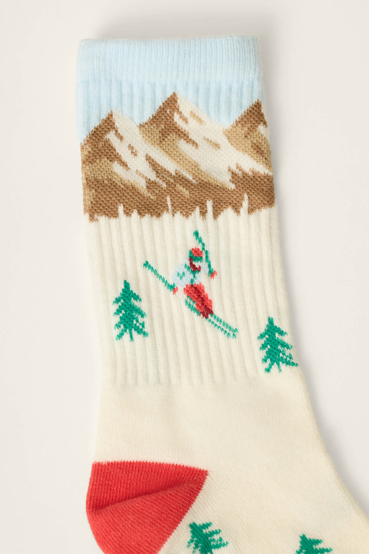 The P.J. Salvage Snowy Mountainscape Crew Socks feature a cream background with a red and green skier, green trees, brown mountains under a blue sky, and a red heel.