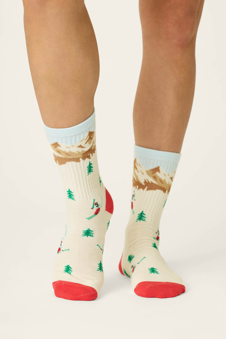 A person wears P.J. Salvage Snowy Mountainscape Crew Socks—cream socks featuring red toes and heels, decorated with green trees, skiers, and brown mountains on a plain white background.
