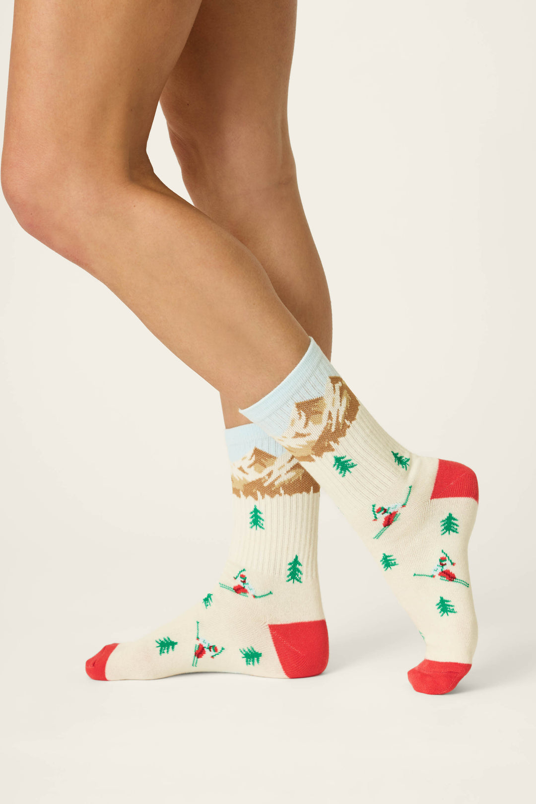 A person wearing P.J. Salvage Snowy Mountainscape Crew Socks—cream socks featuring red toes and heels, adorned with skiers, trees, and mountains—stands with one leg crossed behind the other against a plain background.