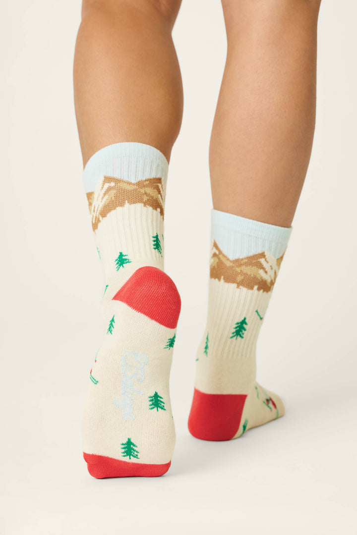 A person is shown from behind wearing the P.J. Salvage Snowy Mountainscape Crew Socks, featuring a cream base with green trees, brown mountains, and red toes and heels in a nature-inspired design against a plain background.