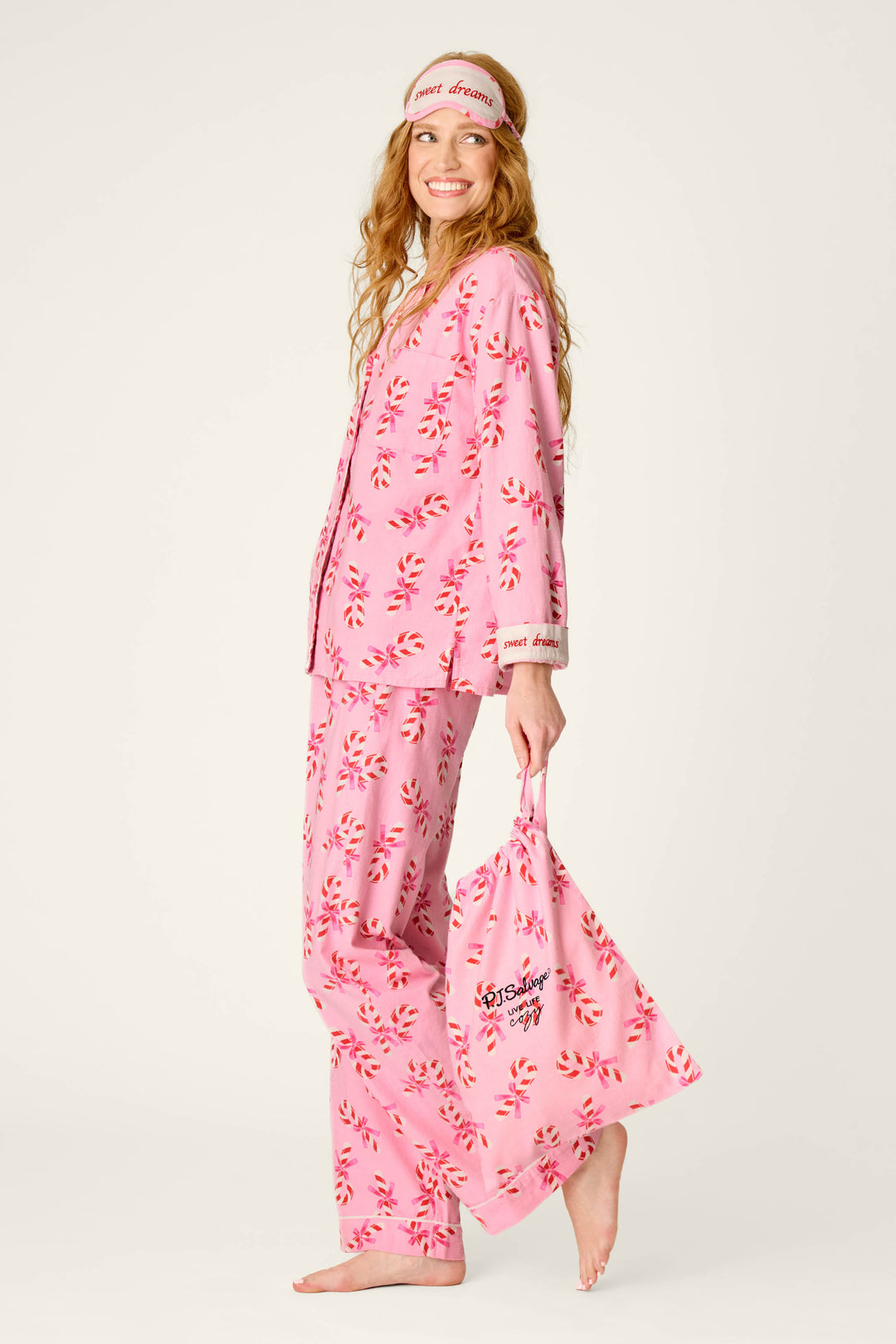 A smiling woman models the P.J. Salvage Sweeter Than Sugar Candy Cane Print 4-Piece PJ Set, featuring pink pajamas with a red and white candy cane pattern, a matching sleep mask, and coordinating tote bag. She is barefoot and stands against a plain background.