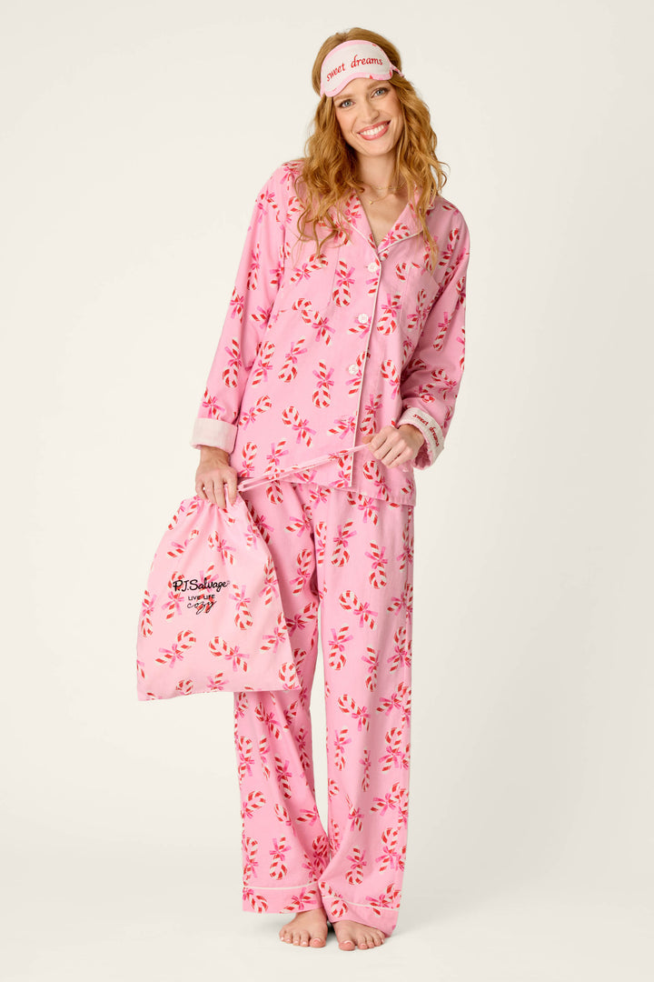 A woman models the P.J. Salvage Sweeter Than Sugar Candy Cane Print 4-Piece PJ Set, featuring pink pajamas with a playful red candy cane pattern. She holds the coordinating bag and wears the matching pink sleep mask embroidered with “sweet dreams,” smiling barefoot against a simple light background.