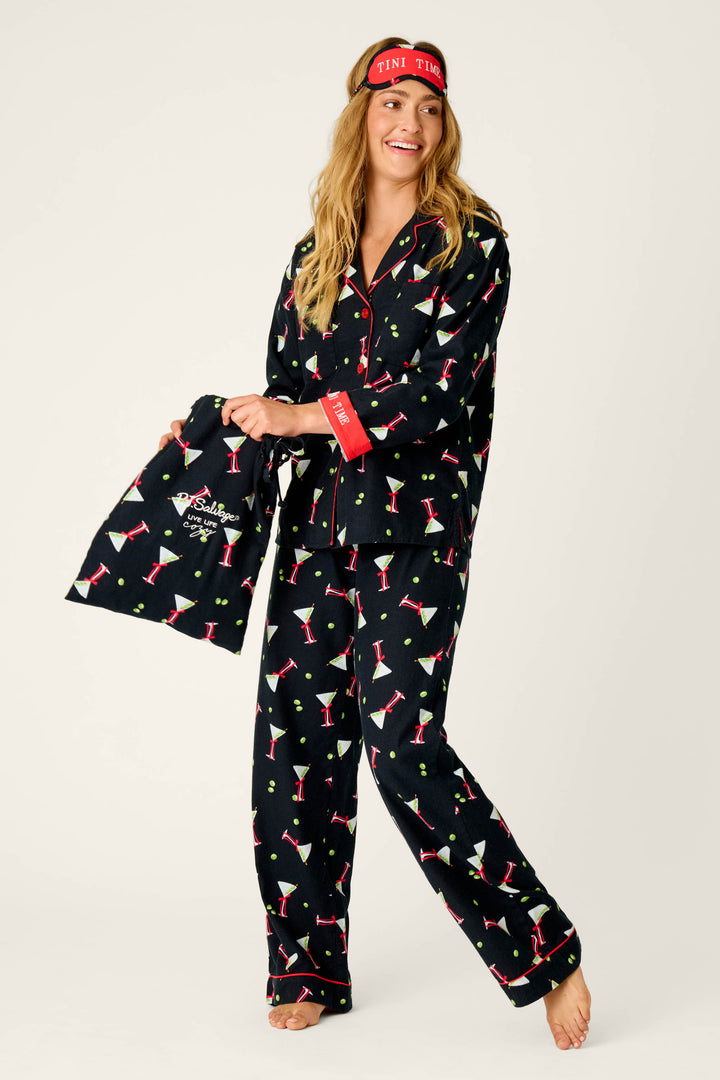 A woman is smiling barefoot against a plain background, wearing the P.J. Salvage Christmas Cocktails Martini Print 4-Piece PJ Set, which features black pajamas decorated with a cocktail glass pattern. She holds a matching bag and wears a red eye mask embroidered with “TINI TIME.”.