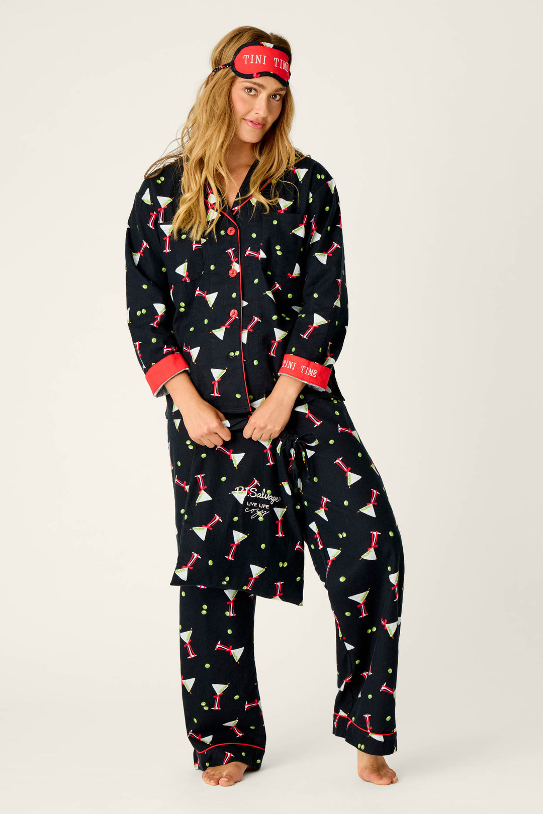 A woman stands barefoot on a plain background, holding the edge of her pajama top. She wears the P.J. Salvage Christmas Cocktails Martini Print 4-Piece PJ Set—black pajamas with a playful martini glass print, red trim, and a coordinating eye mask that reads “TINI TIME.”.