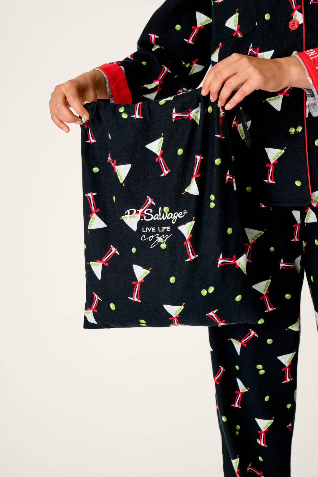 A person wearing the P.J. Salvage Christmas Cocktails Martini Print 4-Piece PJ Set holds a matching drawstring bag that says “PJ Salvage LIVE LIFE Cozy.”.