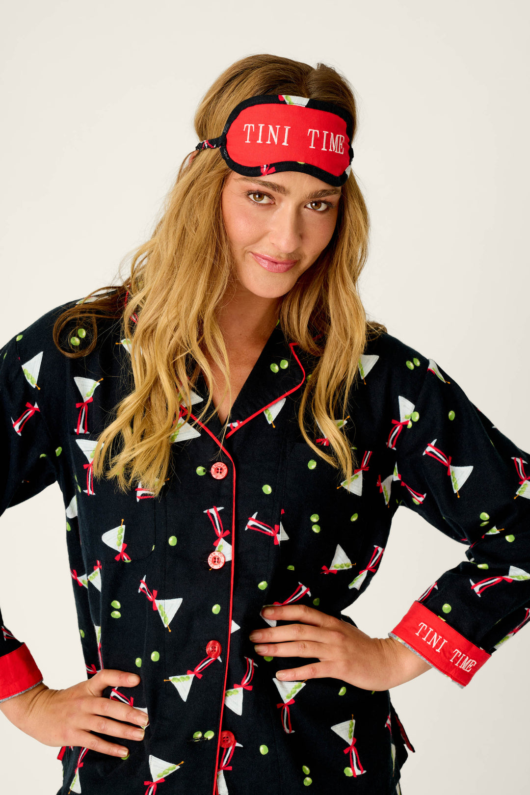A woman smiles with one hand on her hip, dressed in the P.J. Salvage Christmas Cocktails Martini Print 4-Piece PJ Set—featuring dark pajamas with martini glass patterns, red trim, and a matching red sleep mask that reads “TINI TIME”—against a plain background.