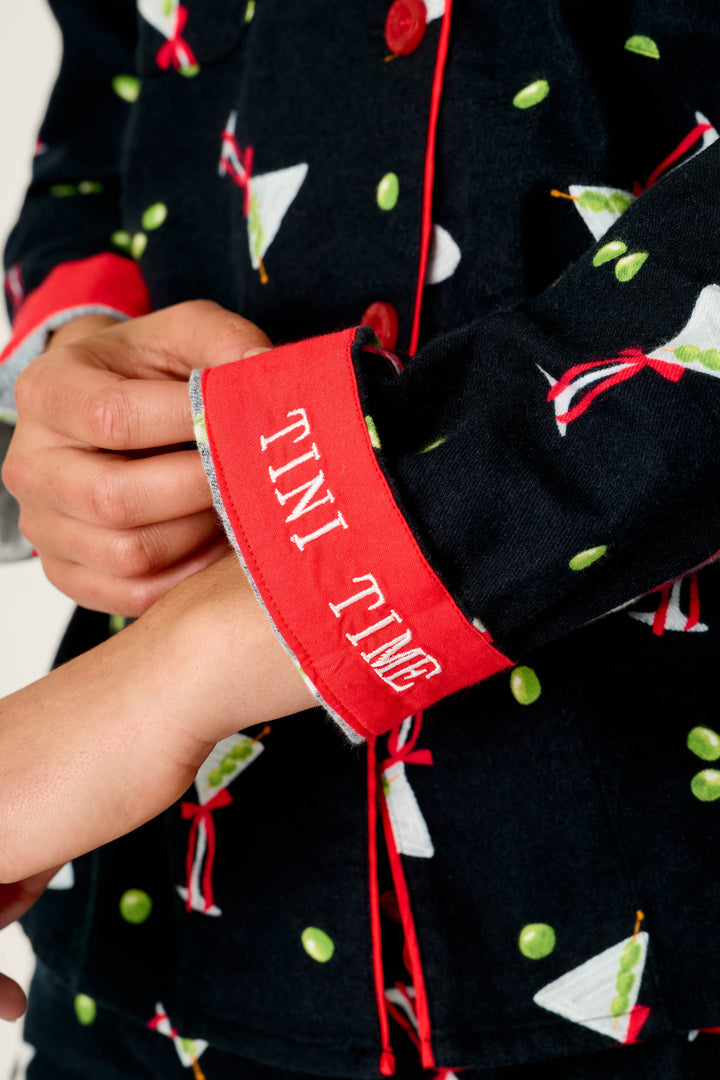 A person adjusts the red cuff of a black sleeve from the P.J. Salvage Christmas Cocktails Martini Print 4-Piece PJ Set, which is decorated with martini glasses and green dots and features “TINI TIME” embroidered in white letters.