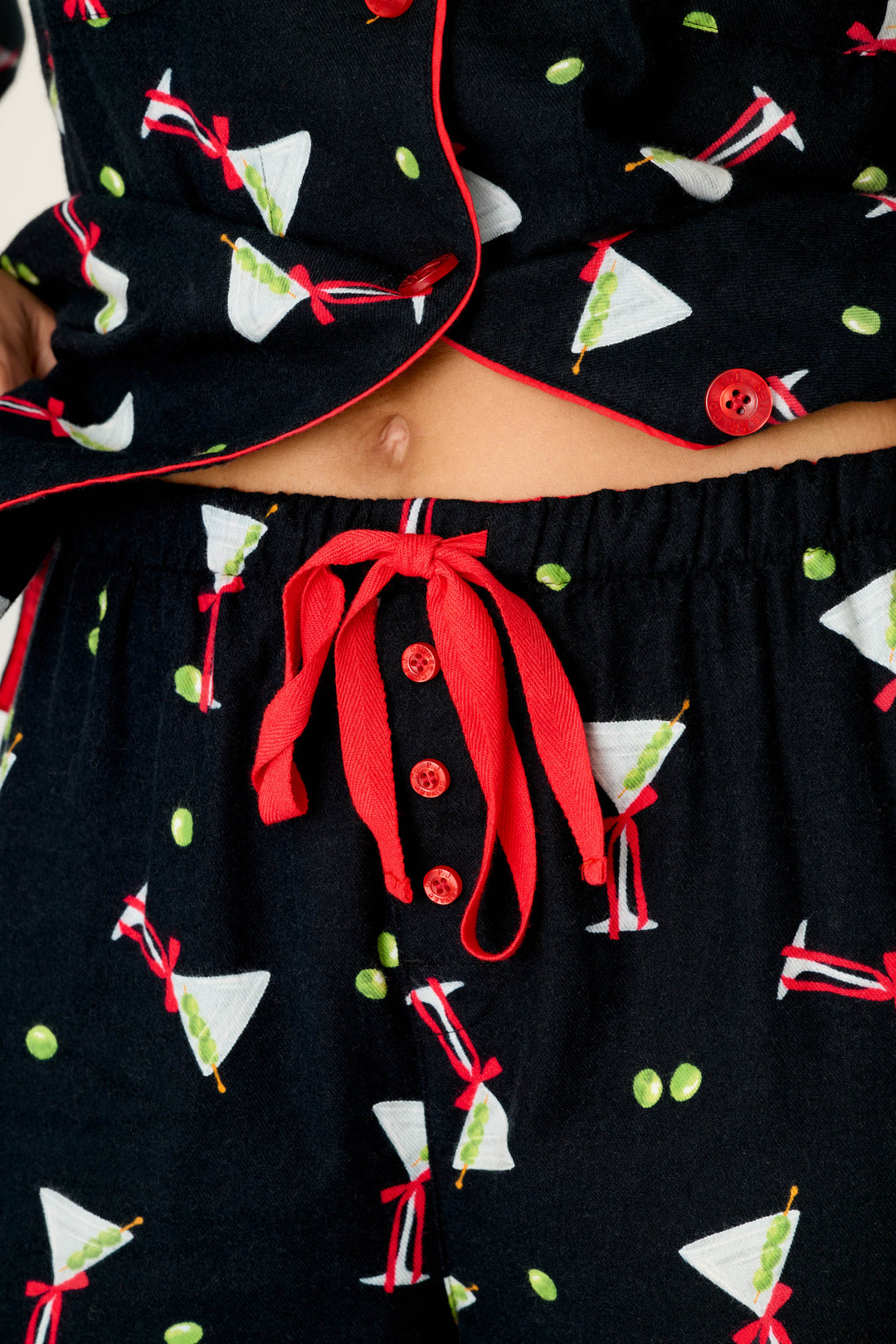 A close-up of a person wearing the P.J. Salvage Christmas Cocktails Martini Print 4-Piece PJ Set, which features black fabric adorned with martini glasses and olives, red buttons, and a red drawstring at the waist; their navel is visible above the waistband.