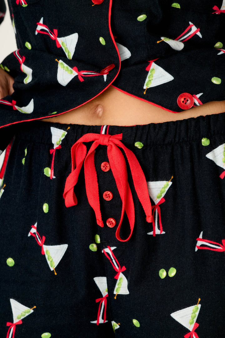 A close-up of a person wearing the P.J. Salvage Christmas Cocktails Martini Print 4-Piece PJ Set, which features black fabric adorned with martini glasses and olives, red buttons, and a red drawstring at the waist; their navel is visible above the waistband.