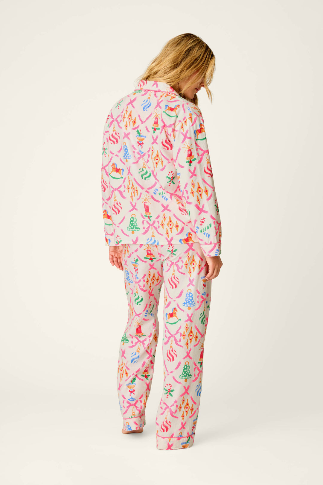 A person with blonde hair stands facing away, wearing the P.J. Salvage Deck the Halls Ornament Print 4-Piece PJ Set, featuring colorful Christmas trees, birds, and festive designs on a white background.