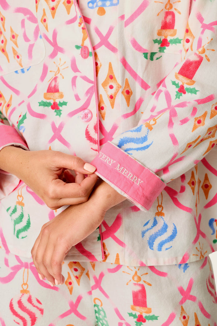 A person wears the P.J. Salvage Deck the Halls Ornament Print 4-Piece PJ Set, featuring festive pink bows and ornaments on the fabric. Their hand pulls back a pink cuff embroidered with “MERRY MERRY” in white letters.