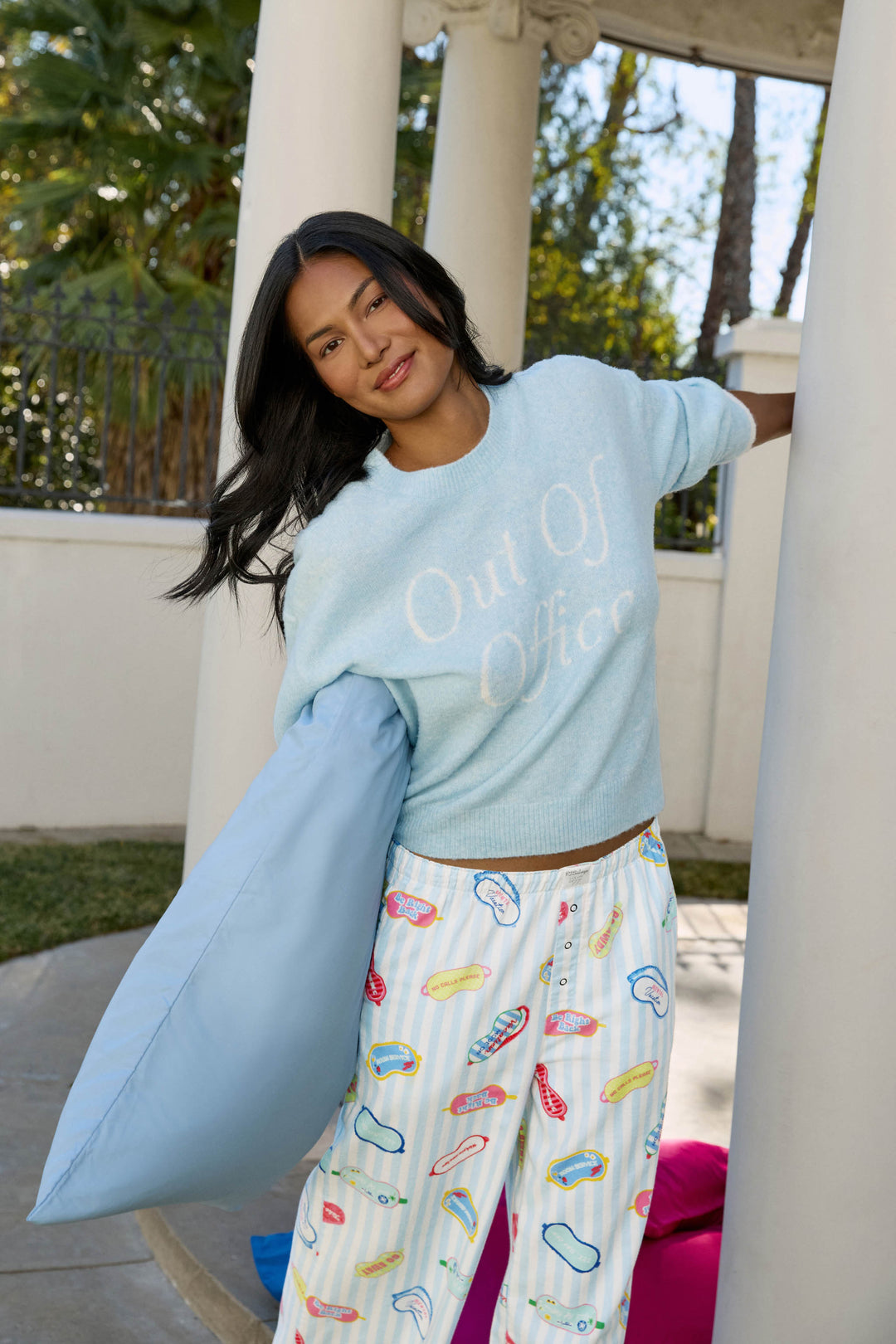 A woman wearing P.J. Salvage pajamas with a colorful lips print and the Out of Office Snuggle Sweater in light blue leans against a column outdoors, holding a blue pillow, with palm trees and a white fence in the background.