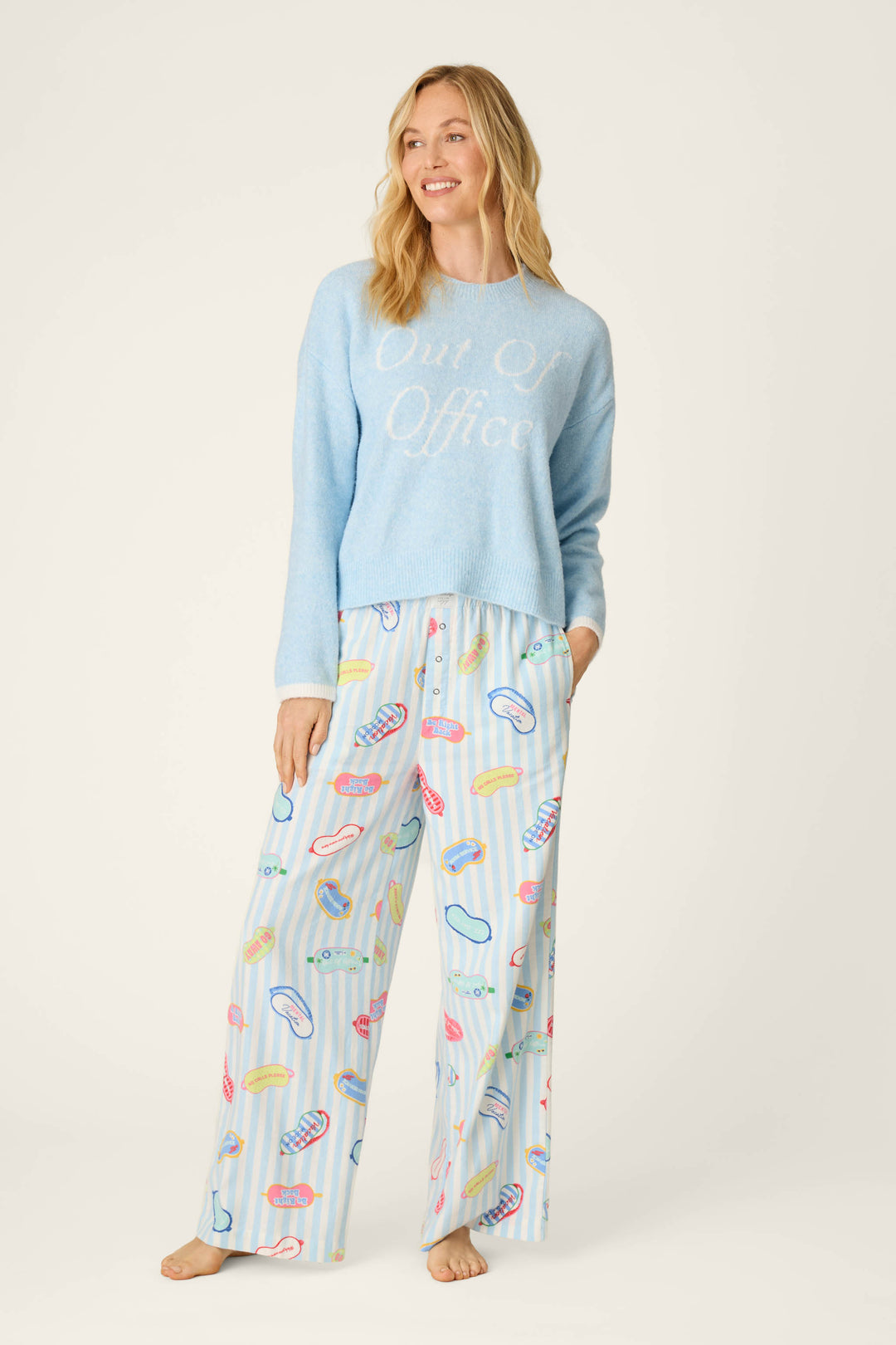 A woman smiles while wearing the P.J. Salvage Out of Office Snuggle Sweater in light blue and pajama pants with a colorful macaron print, standing barefoot against a plain light background.