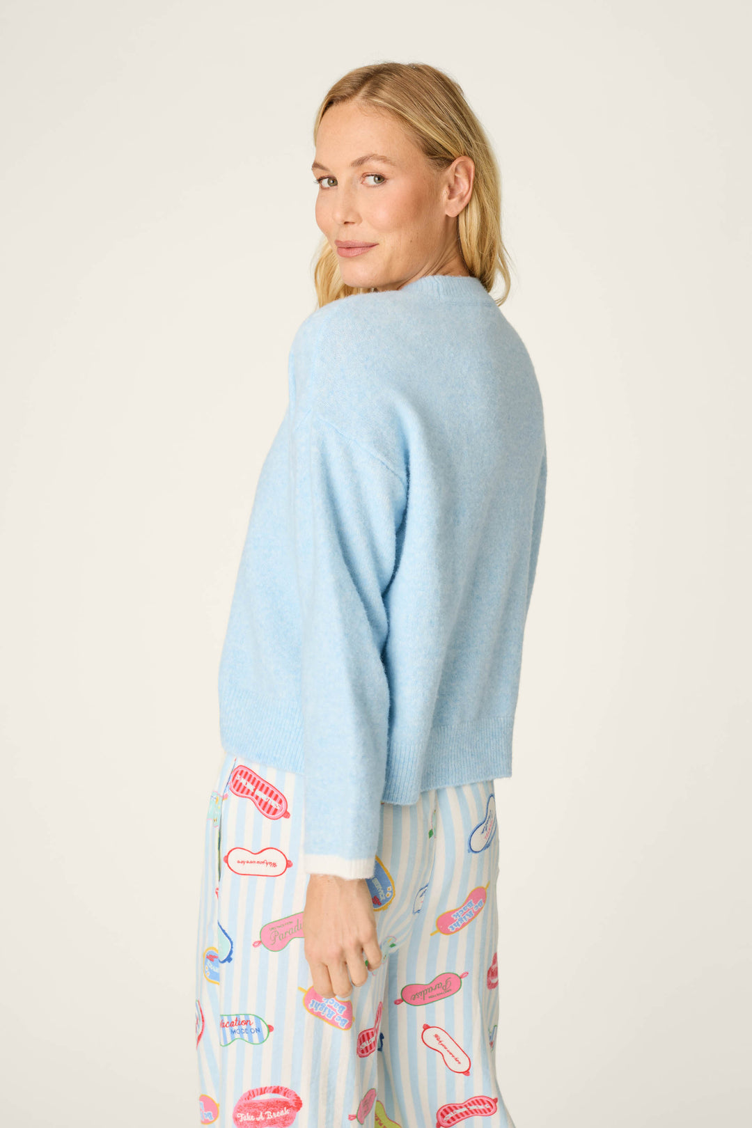 A woman with blonde hair wears the P.J. Salvage Out of Office Snuggle Sweater in light blue and vibrant, tooth-and-toothbrush patterned pants, standing sideways and looking over her shoulder with a slight smile against a plain background.