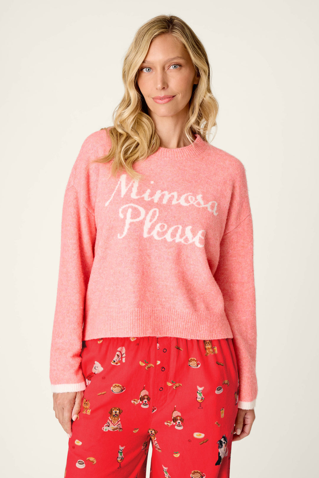 A woman with long blonde hair wears the P.J. Salvage Mimosa Please Snuggle Sweater in pink, paired with red pajama pants featuring various cartoon prints, standing against a plain background.