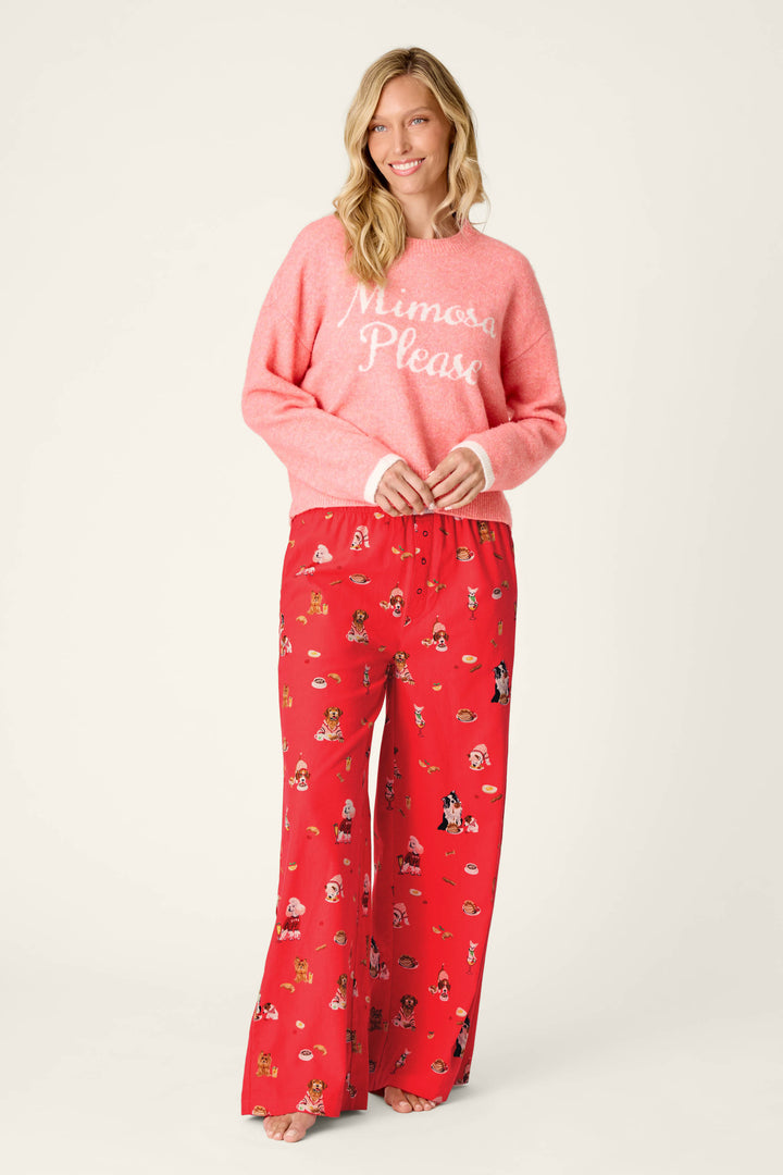 A woman stands barefoot and smiles, wearing the P.J. Salvage Mimosa Please Snuggle Sweater—a fuzzy pink top—paired with loose red pajama pants featuring cartoon characters, set against a plain light background.