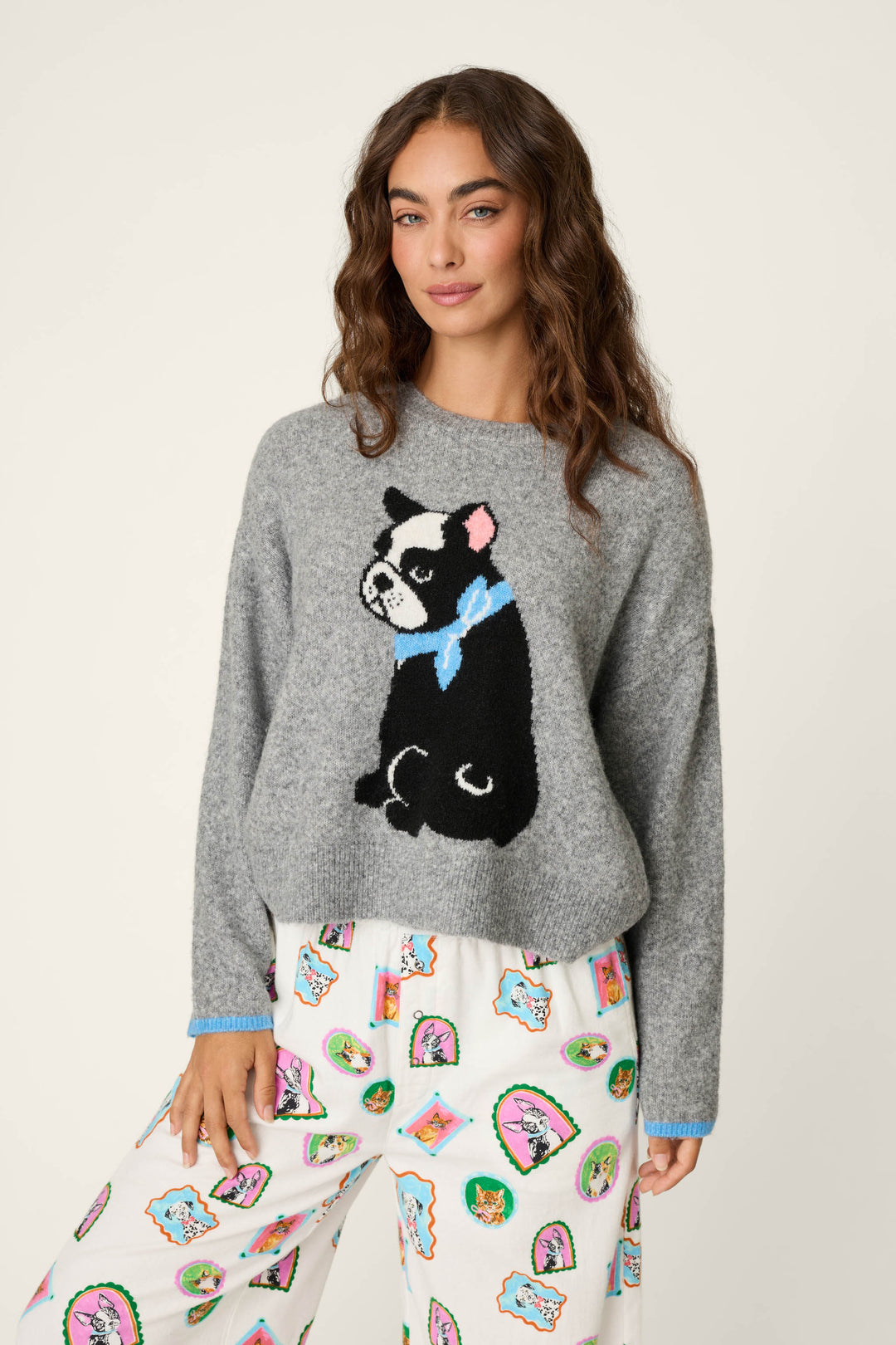 A woman with wavy brown hair wears the P.J. Salvage French Bulldog Snuggle Sweater—a grey sweater featuring a black and white dog with a blue bow—and white pajama pants decorated with colorful cartoon dog faces, standing against a plain background.