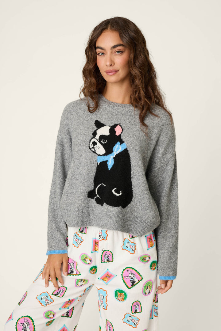 A woman with wavy brown hair wears the P.J. Salvage French Bulldog Snuggle Sweater—a grey sweater featuring a black and white dog with a blue bow—and white pajama pants decorated with colorful cartoon dog faces, standing against a plain background.