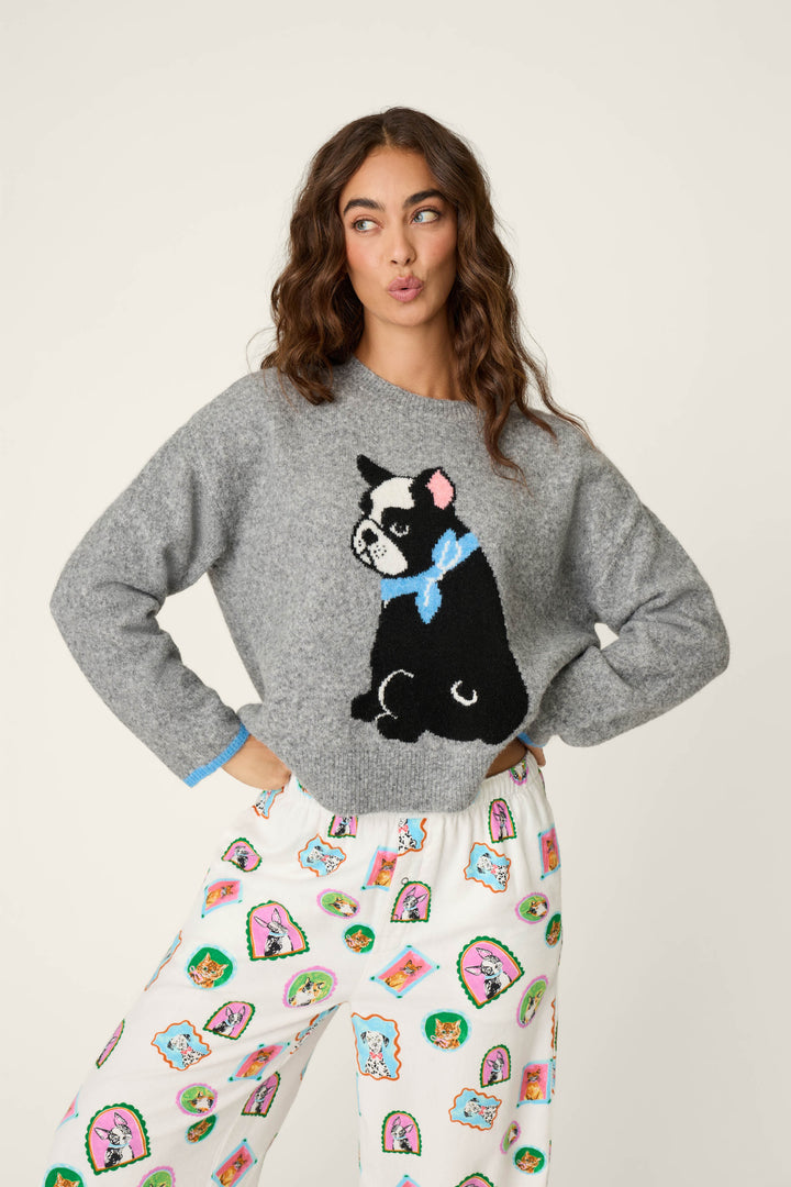 A woman with wavy brown hair stands with her hands on her hips, making a playful pout while wearing the P.J. Salvage French Bulldog Snuggle Sweater and white pajama pants decorated with colorful cartoon faces.