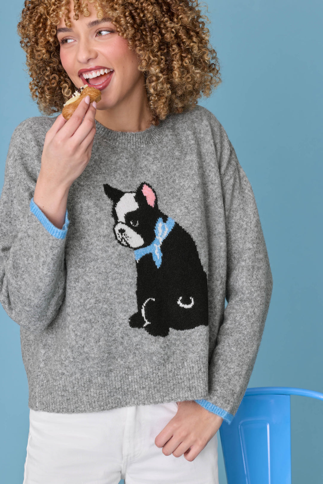 A smiling person with curly hair enjoys a pastry while wearing the P.J. Salvage French Bulldog Snuggle Sweater, which features a black and white dog design and light blue trim, standing against a blue background.