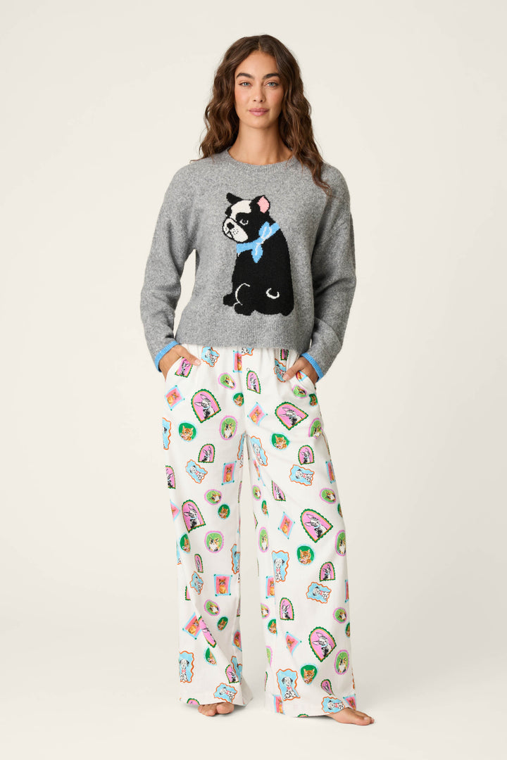 A woman stands barefoot against a plain light background, hands in pockets, wearing the P.J. Salvage French Bulldog Snuggle Sweater— a gray top with a black dog graphic—paired with loose white pajama pants decorated with colorful dog faces and taco illustrations.