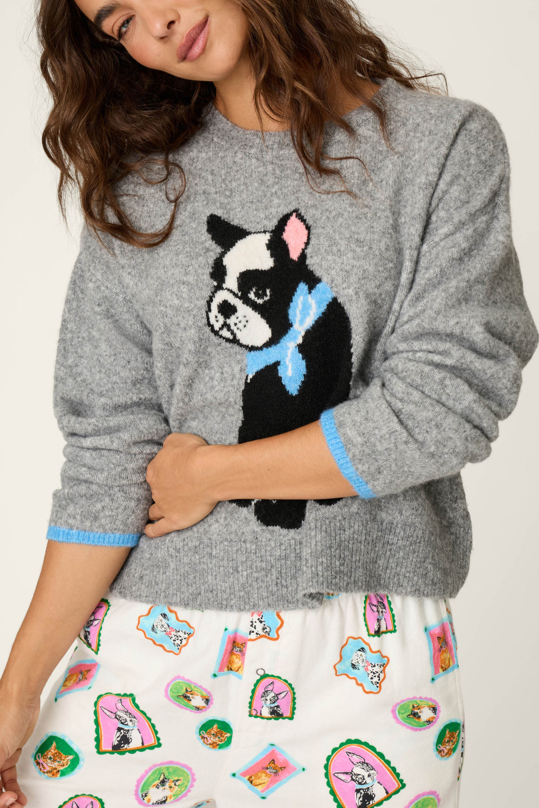 A woman wears the P.J. Salvage French Bulldog Snuggle Sweater, a grey top featuring a cartoon dog design, paired with white pants adorned with colorful cartoon dog illustrations, standing against a plain background.