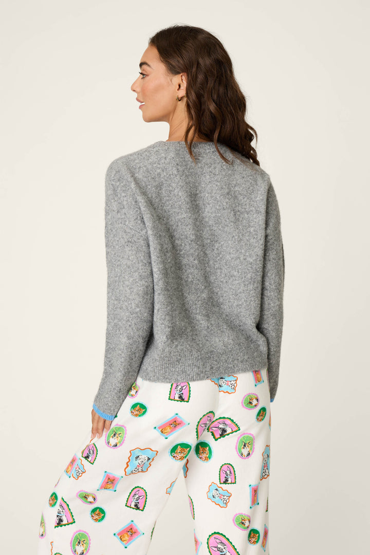 A woman faces away, wearing the P.J. Salvage French Bulldog Snuggle Sweater in gray paired with white pants adorned with colorful cartoon faces and shapes. The background is plain and light-colored.