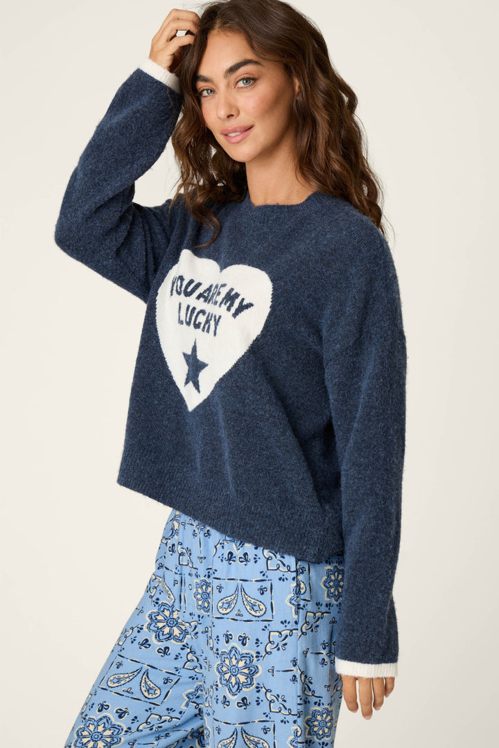 A woman with long brown hair wears the P.J. Salvage You Are My Lucky Star Snuggle Sweater—a blue fuzzy style featuring a white heart and “You are my lucky” text—paired with blue patterned pants, standing against a plain background.