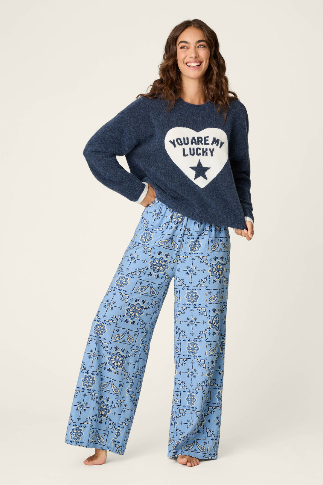 A woman smiles while wearing the P.J. Salvage “You Are My Lucky Star Snuggle Sweater,” a navy top with a white heart graphic, paired with loose blue floral pajama pants, standing barefoot against a plain light background.