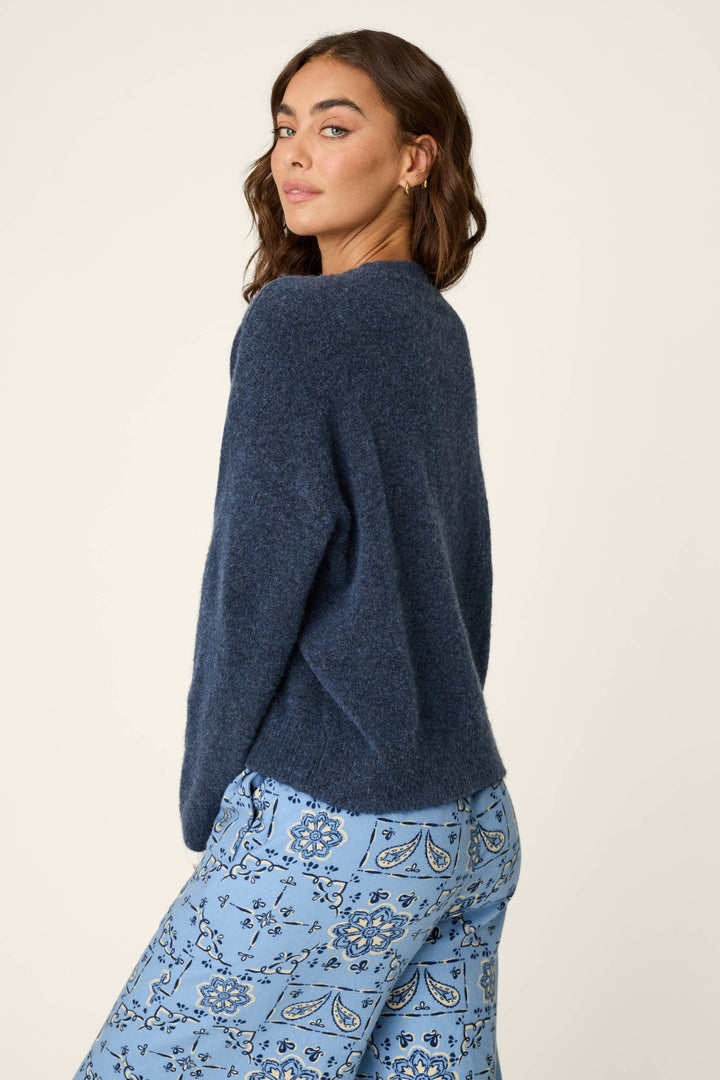 Wearing the P.J. Salvage You Are My Lucky Star Snuggle Sweater, a woman with wavy brown hair poses against an off-white background, glancing over her shoulder in the cozy dark blue top paired with light blue floral patterned pants.