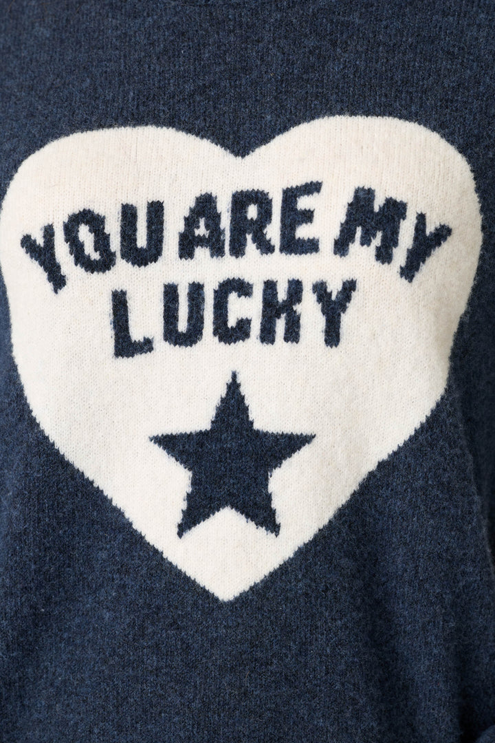 The P.J. Salvage You Are My Lucky Star Snuggle Sweater is a navy blue sweater featuring a large white heart on the front, with “YOU ARE MY LUCKY” written in dark letters above a star shape inside the heart.
