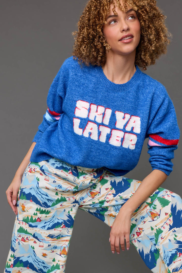 A person with curly hair poses against a gray background wearing the P.J. Salvage Ski Ya Later Snuggle Sweater—a blue top featuring “SKI YA LATER” in bold white and pink letters—and colorful pajama pants decorated with a mountain and camping scene.