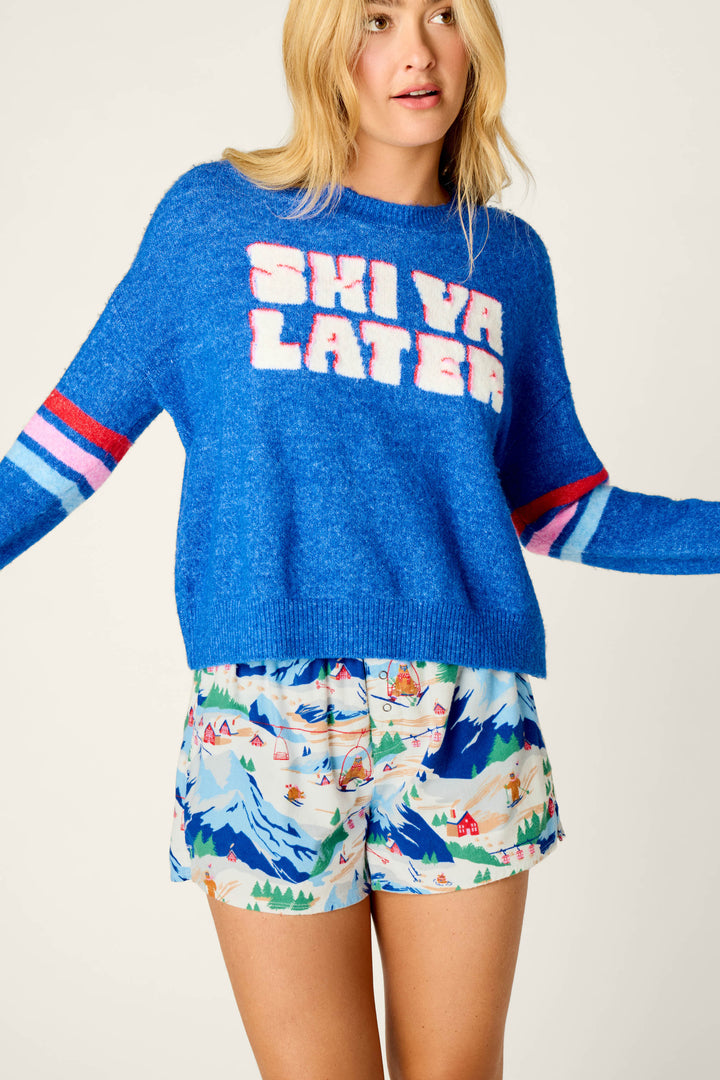 A person wearing the P.J. Salvage Ski Ya Later Snuggle Sweater in bright blue, paired with colorful ski mountain print shorts, stands against a plain background.
