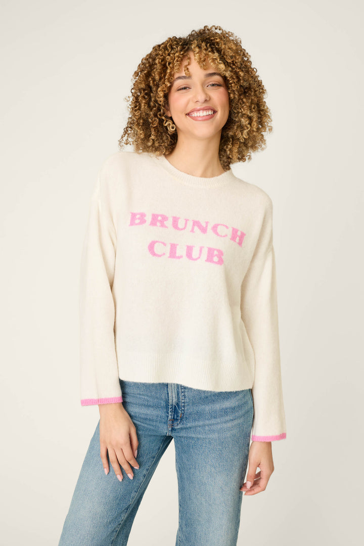 A smiling person with curly hair is wearing the Brunch Club Snuggle Sweater by P.J. Salvage— a cream sweater featuring pink "BRUNCH CLUB" lettering—paired with blue jeans, standing against a plain background.