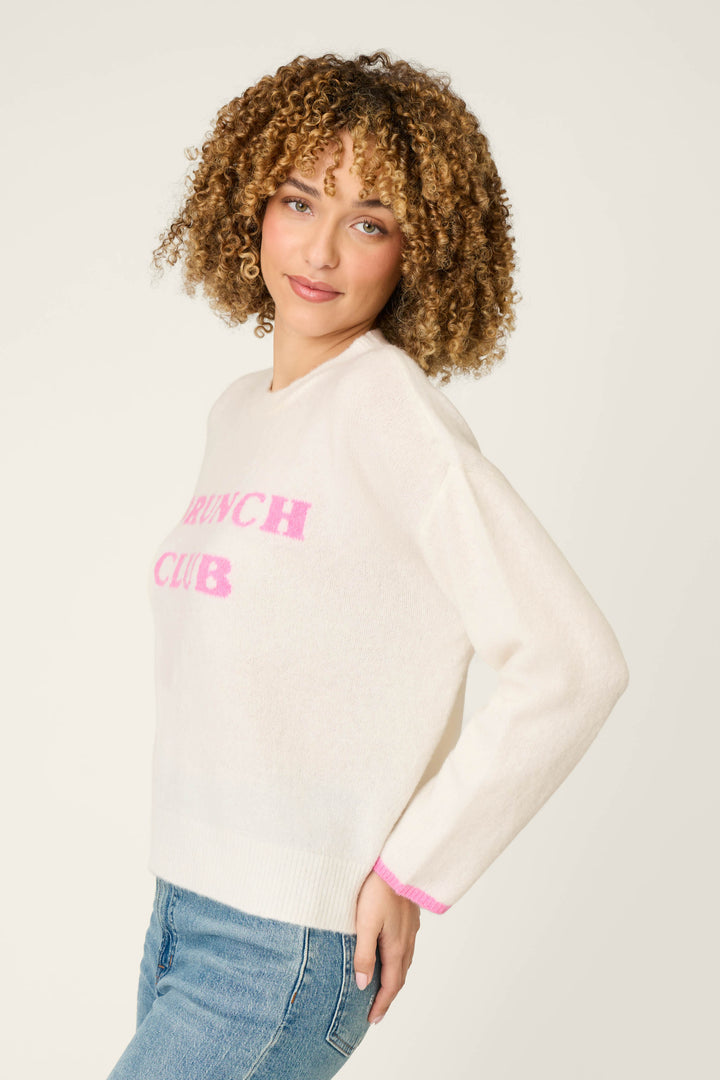 A woman with curly hair wears the P.J. Salvage Brunch Club Snuggle Sweater—a white pullover featuring pink "BRUNCH CLUB" text—paired with blue jeans, posing against a light background.