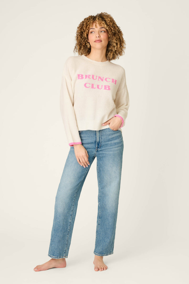 A person with curly hair wears the P.J. Salvage Brunch Club Snuggle Sweater, featuring "BRUNCH CLUB" in pink letters, paired with blue jeans and stands barefoot against a plain light background.
