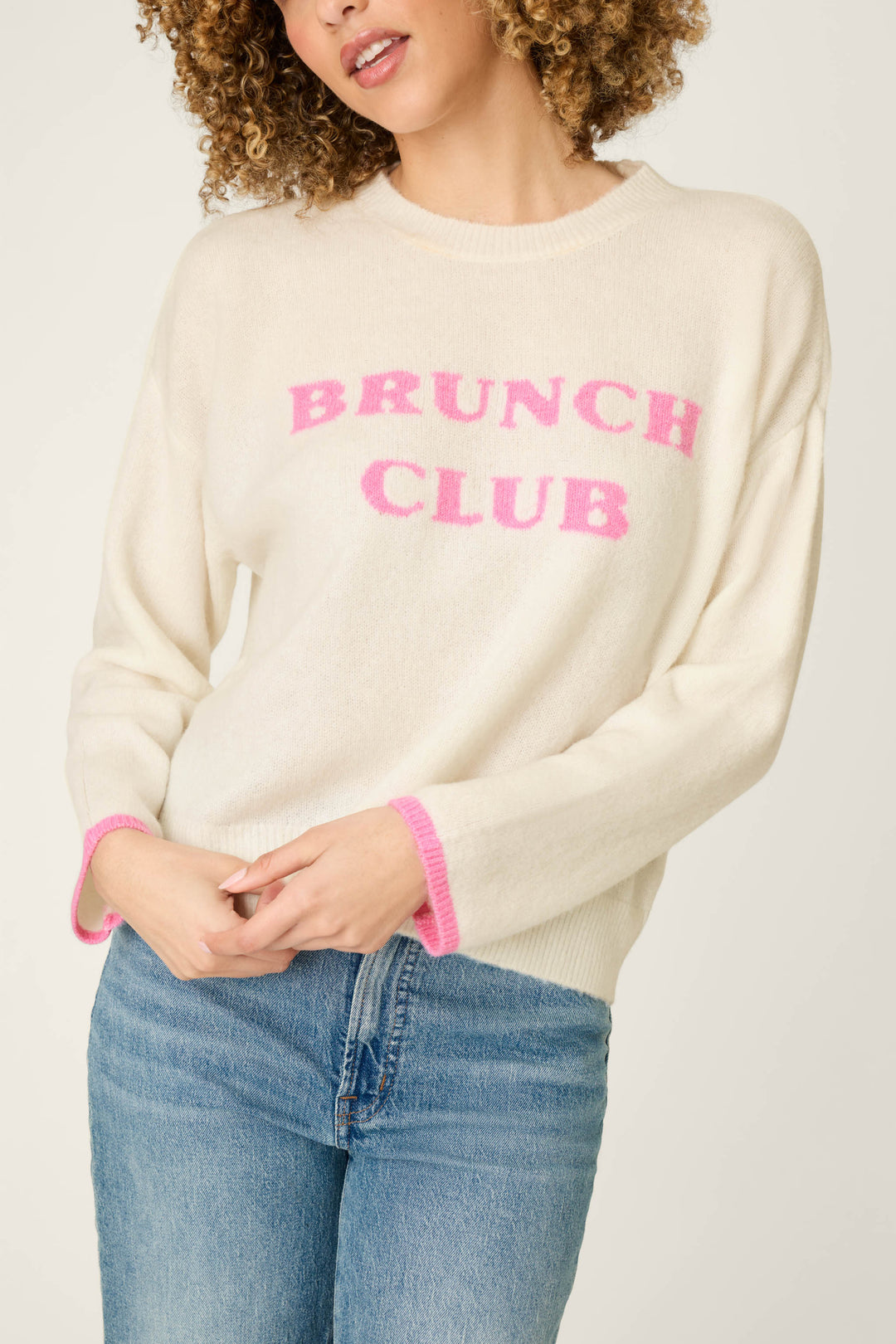 A woman wears the P.J. Salvage Brunch Club Snuggle Sweater, a cream sweater featuring pink trim and “BRUNCH CLUB” in pink lettering on the front, styled with blue jeans. Her face is partially visible and her curly hair is worn down.