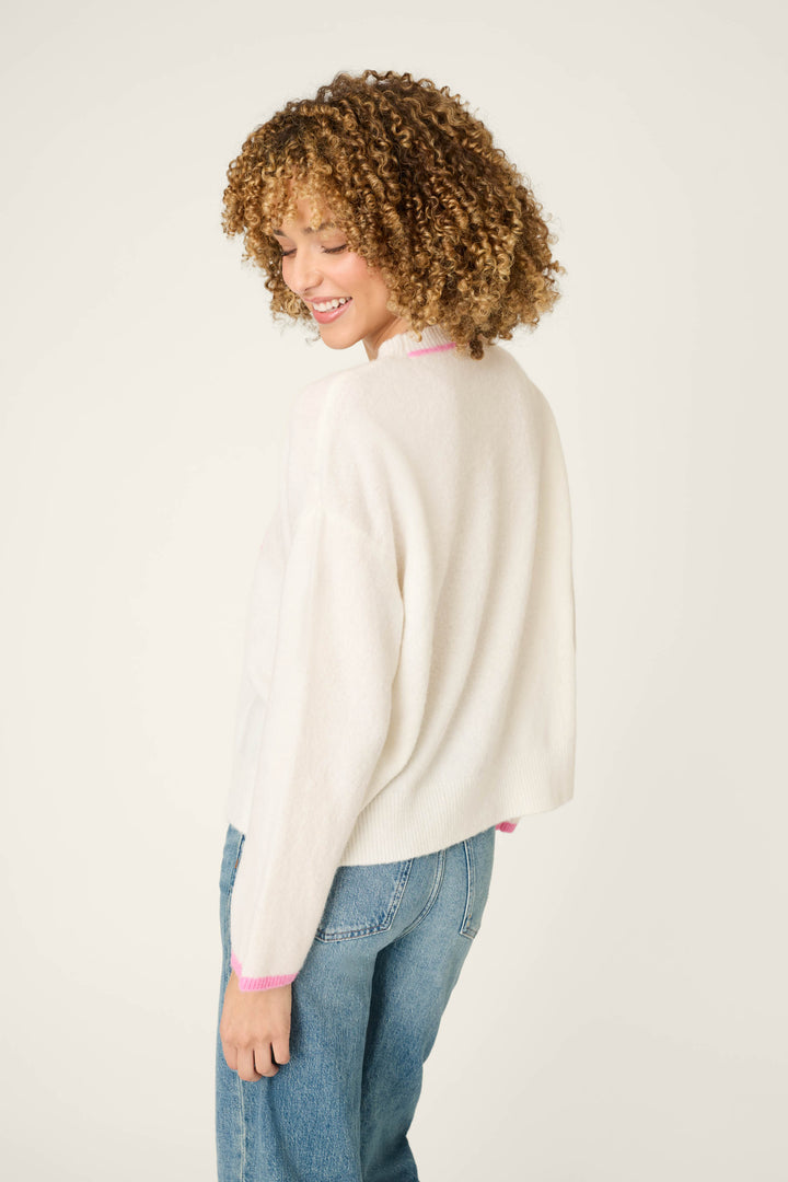 Wearing the Brunch Club Snuggle Sweater by P.J. Salvage, a person with curly hair stands sideways and smiles while looking down, pairing the white sweater with pink trim with blue jeans.