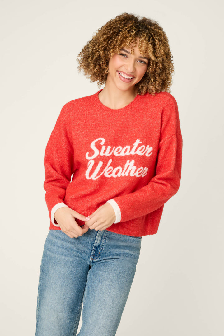 A smiling person with curly hair is wearing the P.J. Salvage Sweater Weather Snuggle Sweater in red, featuring “Sweater Weather” written in white, paired with light blue jeans and standing against a plain light background.