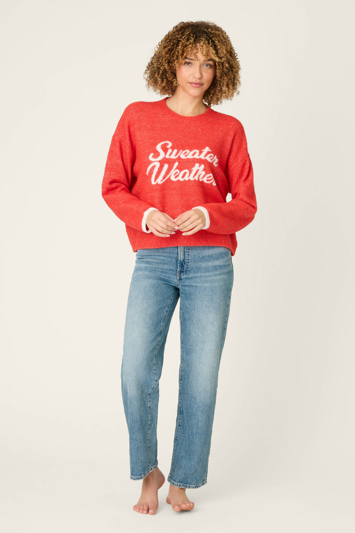 A person with curly hair stands barefoot against a plain light background, wearing blue jeans and the P.J. Salvage Sweater Weather Snuggle Sweater in red.