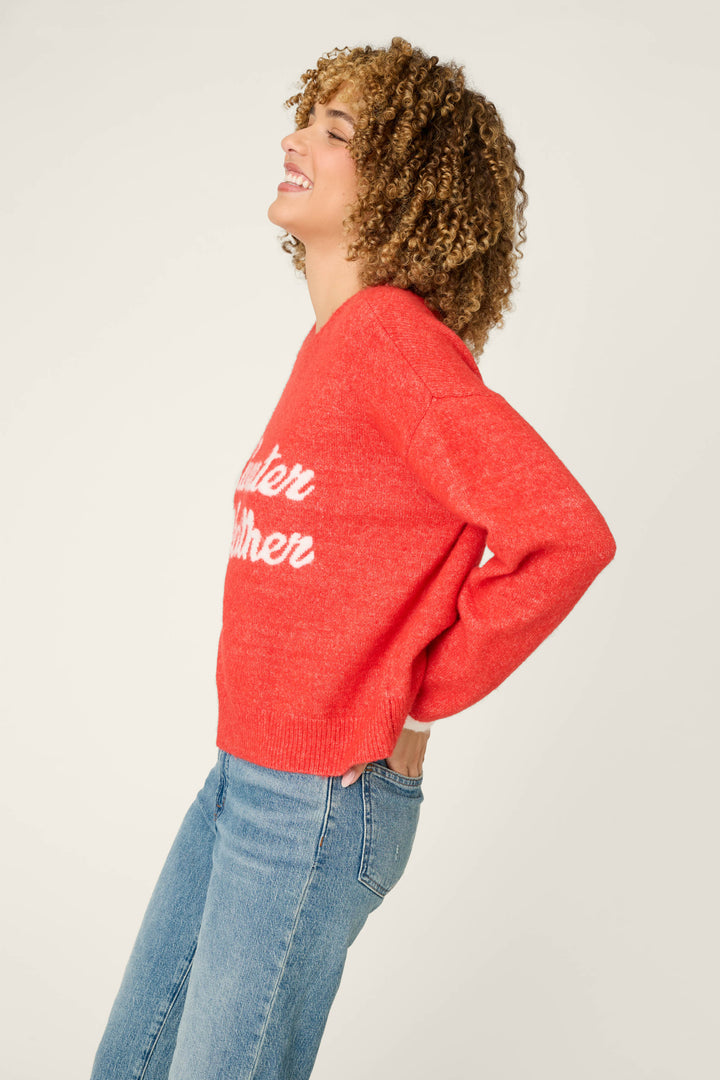 A person with curly hair smiles while wearing the P.J. Salvage Sweater Weather Snuggle Sweater—a red sweater with white text—paired with blue jeans, posing in a side profile against a plain light background.