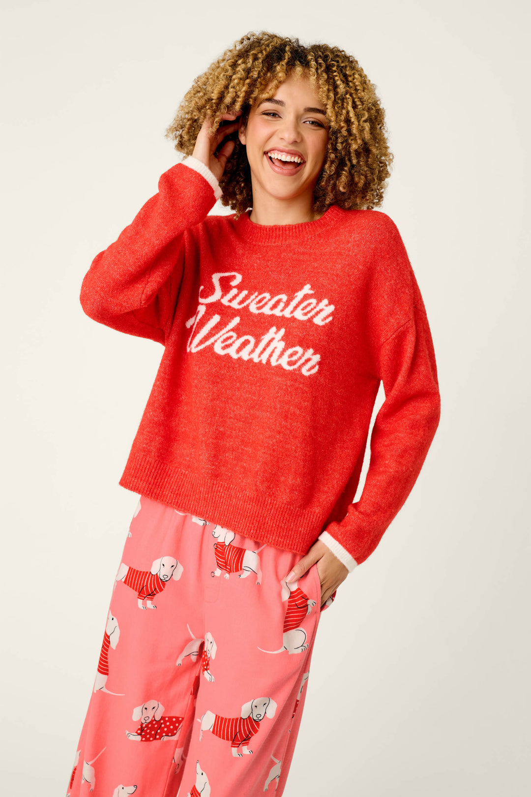 A smiling person wearing a vibrant red P.J. Salvage Sweater Weather Snuggle Sweater and pink pajama pants with dog prints, standing against a plain white background.