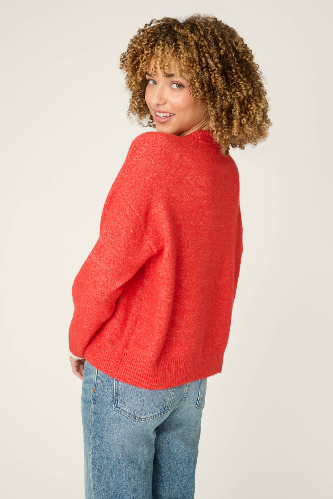 A woman with curly hair wears the P.J. Salvage Sweater Weather Snuggle Sweater in vibrant red and blue jeans, turning slightly to smile over her shoulder at the camera against a plain light background.