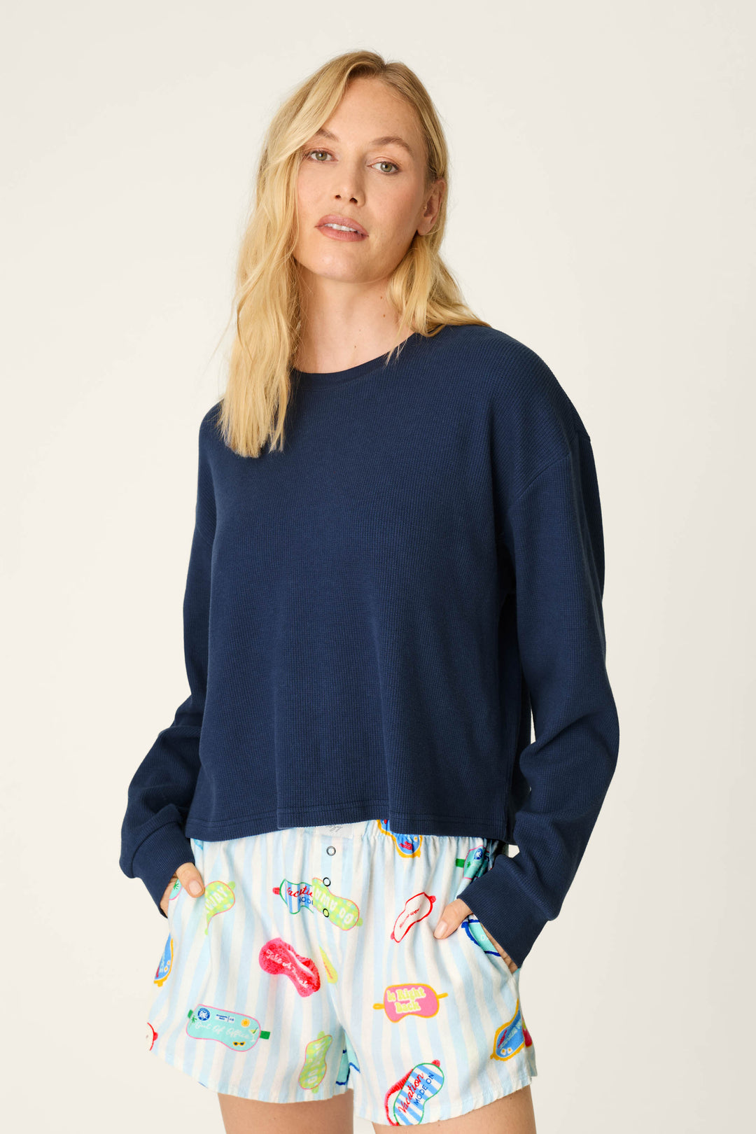 A woman with blonde hair wears the P.J. Salvage Blue Night Long Sleeve Top and light blue shorts with colorful graphic prints, posing with her hands in her pockets against a plain background.