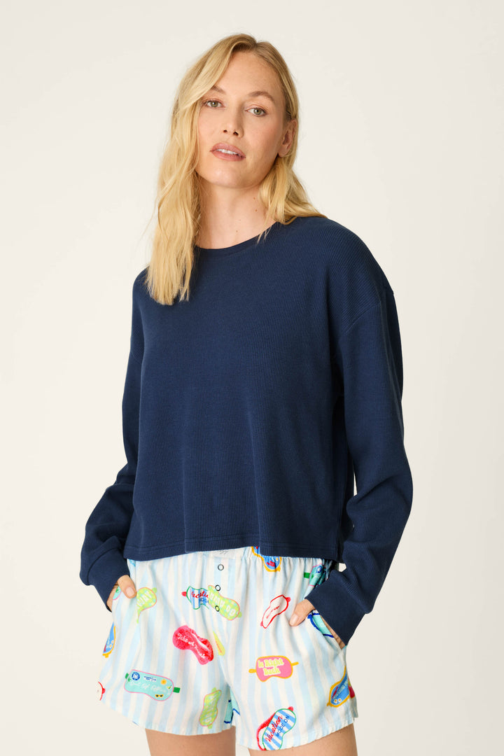 A woman with blonde hair wears the P.J. Salvage Blue Night Long Sleeve Top and light blue shorts with colorful graphic prints, posing with her hands in her pockets against a plain background.