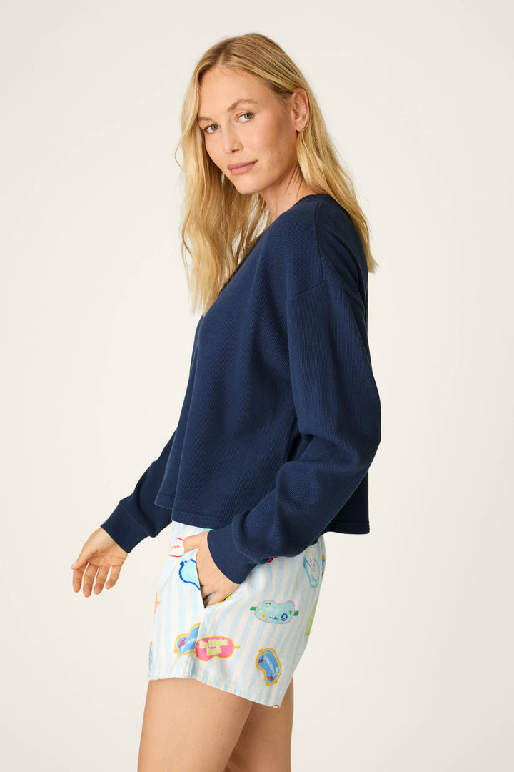 A woman with long blonde hair models the P.J. Salvage Blue Night Long Sleeve Top, paired with white shorts featuring colorful abstract patterns. She stands sideways with one hand in her pocket, looking at the camera against a plain background.