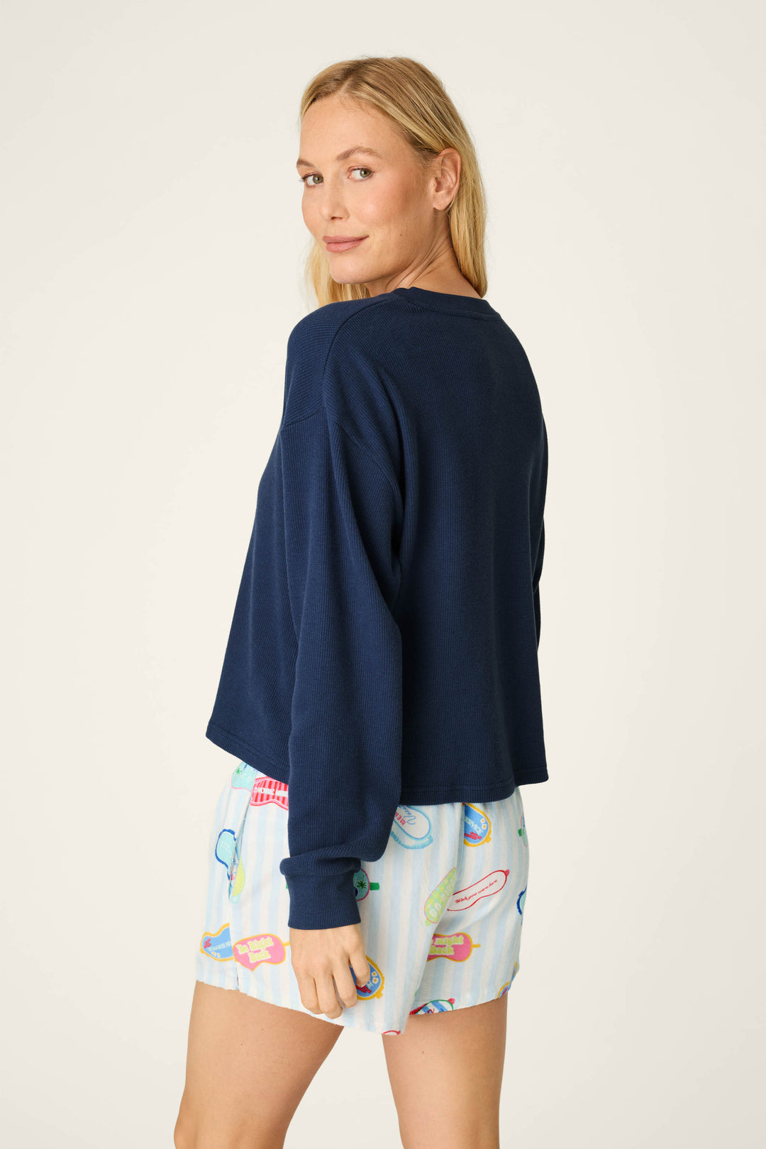 A woman with long blonde hair wears the P.J. Salvage Blue Night Long Sleeve Top and colorful pajama shorts featuring tennis racket and trophy designs, standing and looking back over her shoulder against a plain background.