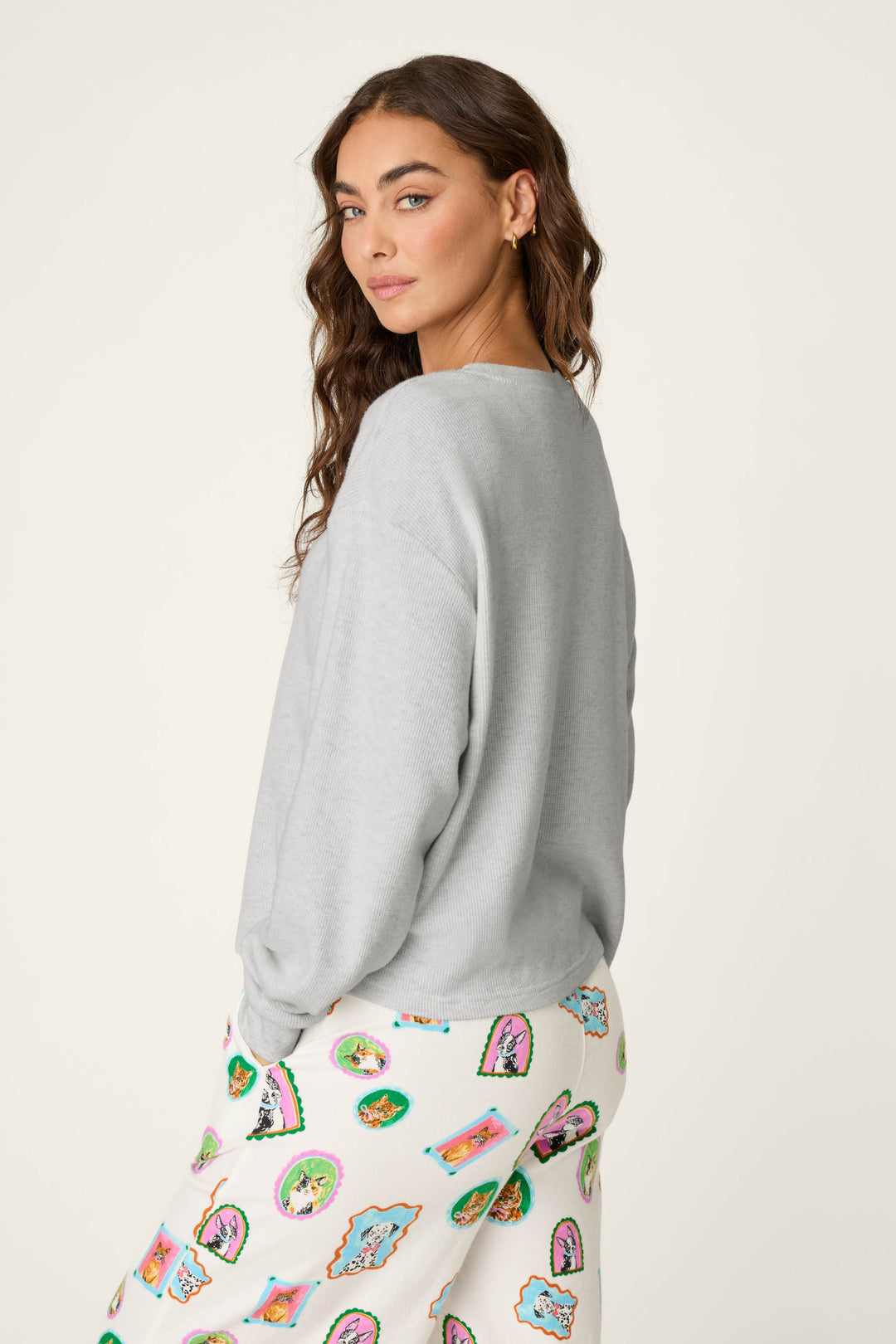 A woman with long wavy brown hair wears the P.J. Salvage Heather Grey Long Sleeve Top and a white skirt with colorful cartoon-style faces, standing against a plain light background and looking over her shoulder.