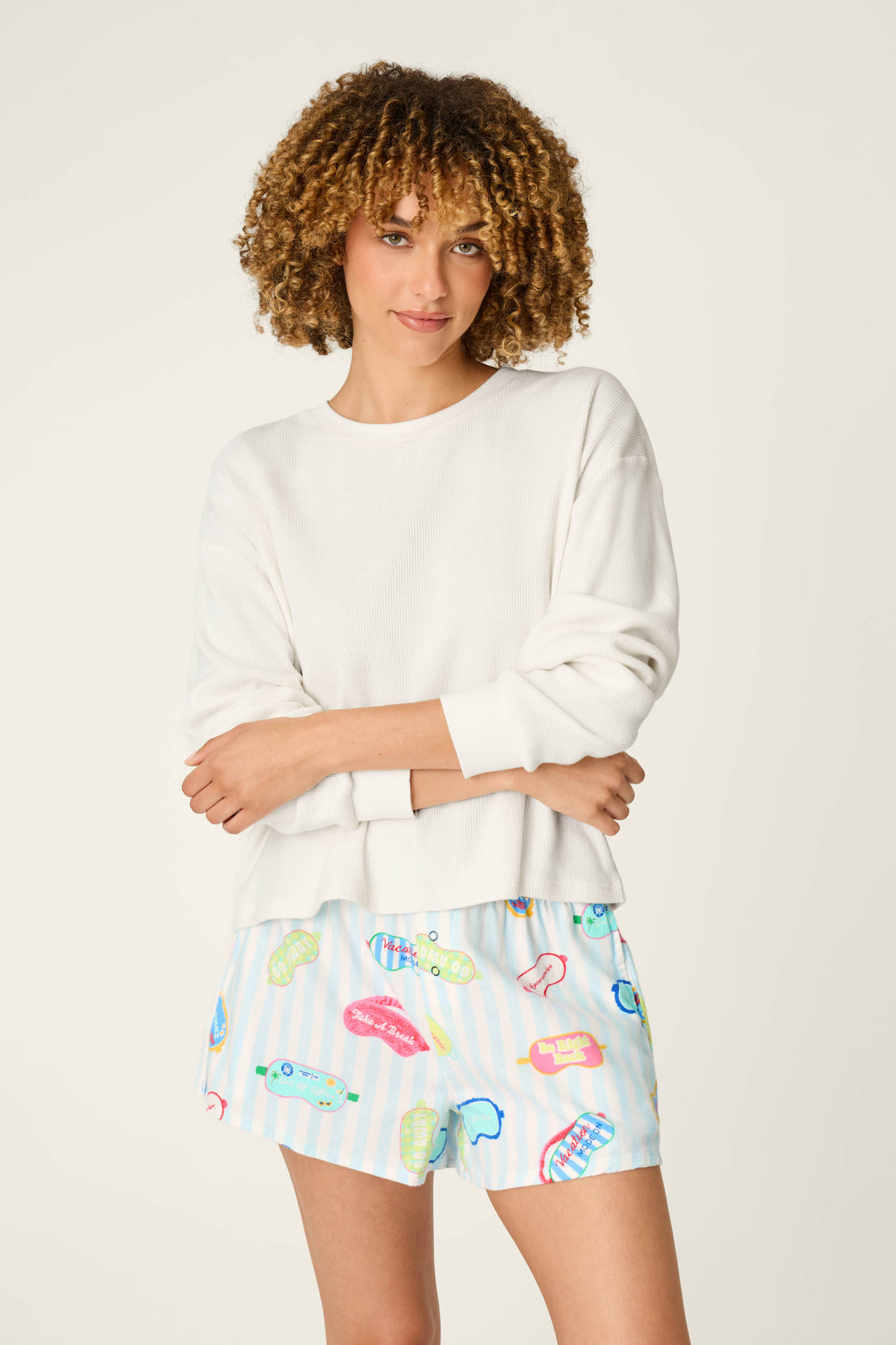 A woman with curly hair stands against a plain light background, arms crossed and smiling slightly, wearing the P.J. Salvage White Long Sleeve Top and colorful printed shorts.