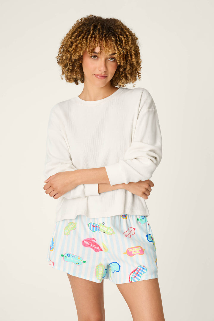 A woman with curly hair stands against a plain light background, arms crossed and smiling slightly, wearing the P.J. Salvage White Long Sleeve Top and colorful printed shorts.